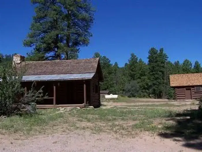 An exterior view of a historic cabin surrounded by trees on a blue sky day. A separate building in the background contains toilets and showers.