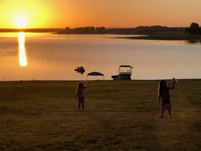 Two children stand near the shore of a lake at sunset