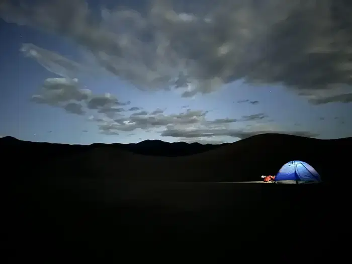 Nighttime. Glowing tent on dark sand dunes. A few stars and clouds in the sky.